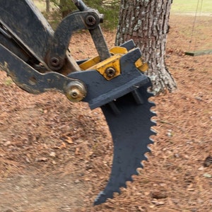 Op de afbeelding: Een close-up van een zwarte graafmachine-aanbouwdeel met een gekarteld blad. De aanbouwdeel is ontworpen voor het graven en opruimen van bomen en struiken.