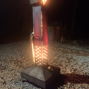 Puede incluir: Una chimenea exterior alta de metal con un cuerpo rectangular y una base cuadrada. La chimenea tiene un brillo rojo y una serie de recortes en forma de media luna. La parte superior presenta las letras "TR" en naranja brillante.