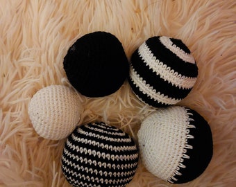 May include: Five crocheted balls in black and white. The balls are arranged in a circle on a fluffy white surface.