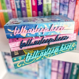 May include: Three decorative book boxes stacked on top of each other, each with the phrase "fell asleep here" in a cursive font. The top box is blue with a bicycle design, the middle is pink, and the bottom is teal. Books are visible in the background.