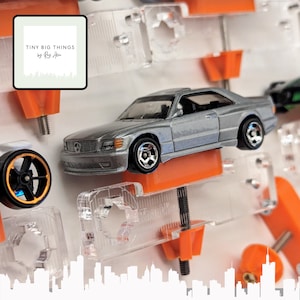 May include: A silver Mercedes-Benz coupe model car is displayed on an orange stand. The car has black wheels and a detailed front grille. The image includes the text "TINY BIG THINGS by Ray Allen" in a white square with a cityscape outline.