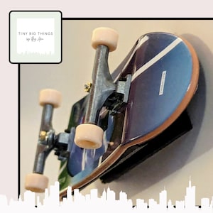 May include: A miniature skateboard with a blue and white deck, black trucks, and beige wheels. The skateboard is mounted on a wall with a white background. The text "TINY BIG THINGS by Rex Allen" is visible in the top left corner of the image.