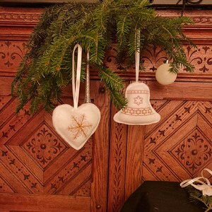 Könnte beinhalten: Weihnachtsschmuck, der an einem Kiefernzweig hängt. Ein weißer herzförmiger Schmuck mit einem goldenen Schneeflockenmuster und ein weißer glockenförmiger Schmuck mit einem goldenen Sternmuster. Eine weiße Kugel ist ebenfalls sichtbar.