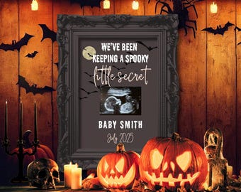 Puede incluir: Anuncio de embarazo con temática de Halloween con una ecografía enmarcada. El marco dice "We've been keeping a spooky little secret" con el nombre del bebé y la fecha de nacimiento. Dos calabazas talladas y una calavera están sobre una mesa de madera.