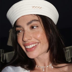 May include: A white pillbox-style hat with a structured, ribbed texture and a small, embroidered cross-stitch pattern in a light tan thread on the front. The hat features a clean, rounded silhouette that sits atop the head, showcasing a simple design.