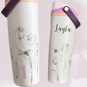 May include: Two white Owala water bottles with floral and bee designs. One bottle has the name "Layla" in script. The bottles have purple handles and lids with pink and peach accents. The Owala logo is visible.