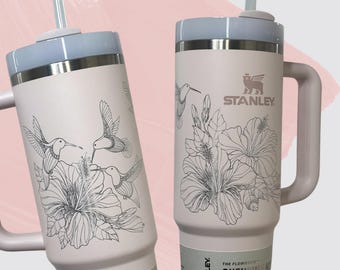 May include: Two light pink Stanley tumblers with a light blue lid and handle. The tumblers feature a black and white hummingbird and floral design. The Stanley logo is visible on one of the tumblers.