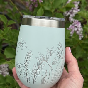 Peut inclure: Un gobelet isotherme bleu clair avec un bord et un couvercle argent&eacute;s. Le gobelet pr&eacute;sente un motif floral gris. La marque "YETI" est visible. Le gobelet est tenu devant un fond de feuilles vertes et de fleurs violettes.