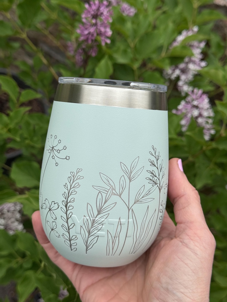 Peut inclure: Un gobelet isotherme bleu clair avec un bord et un couvercle argent&eacute;s. Le gobelet pr&eacute;sente un motif floral gris. La marque "YETI" est visible. Le gobelet est tenu devant un fond de feuilles vertes et de fleurs violettes.
