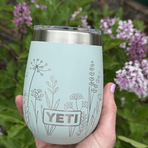 Peut inclure: Un gobelet YETI vert menthe avec un bord et un couvercle argent&eacute;s. Le gobelet pr&eacute;sente un motif floral argent&eacute; et le logo YETI. Le gobelet est tenu dans une main, avec un fond de feuillage vert et de fleurs violettes.