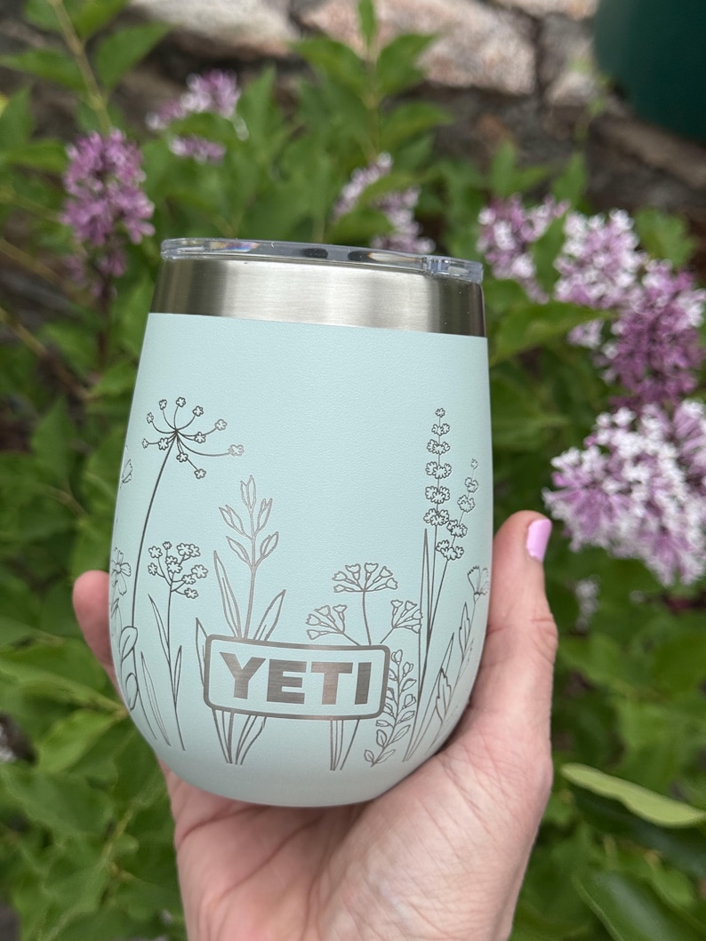 Peut inclure: Un gobelet YETI vert menthe avec un bord et un couvercle argent&eacute;s. Le gobelet pr&eacute;sente un motif floral argent&eacute; et le logo YETI. Le gobelet est tenu dans une main, avec un fond de feuillage vert et de fleurs violettes.