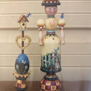May include: Two hand-painted wooden figurines, one depicting a woman with a beehive hat and the other a birdhouse on a post. Both figurines are decorated with colorful details and whimsical designs.