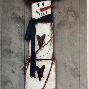 May include: A wooden snowman decoration with a black top hat, blue and white scarf, and red nose. The snowman is made of three stacked wooden blocks with brown hearts and twigs attached.