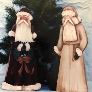 Puede incluir: Dos figuras de madera de Papá Noel. Uno viste un abrigo burdeos oscuro con una corona y un lazo. El otro lleva una túnica beige claro y sostiene un saco negro. Ambos tienen largas barbas blancas y sombreros.