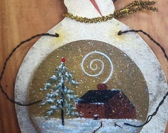 May include: Hand-painted snowman ornament with a winter scene inside. The snowman has a brown hat, a red nose, and a gold tinsel scarf. The scene inside features a house, a snow-covered tree, and a swirling smoke effect.