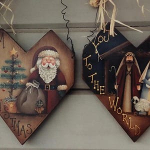 Puede incluir: Dos adornos navideños de madera en forma de corazón. Uno presenta a Papá Noel con un saco y un árbol de Navidad, el otro representa la escena de la Natividad. Las palabras "Merry Christmas" y "You to the World" están pintadas en los adornos.
