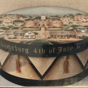 Puede incluir: Una caja redonda de madera con una escena pintada de una ciudad en la parte superior. La caja está decorada con un borde negro y dorado y las palabras "Williamsburg 4th of July 1776".
