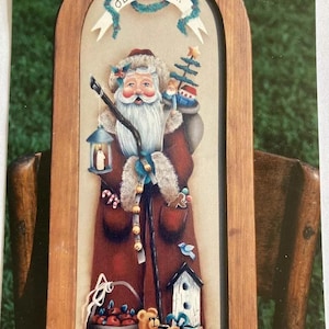 Puede incluir: Una pintura enmarcada en madera de Santa Claus con barba blanca y traje rojo. Lleva un saco de regalos y un bastón. En la escena hay un árbol pequeño, una casita para pájaros, una cesta de manzanas y un osito de peluche. El marco tiene las palabras "Olde St Nick" escritas.