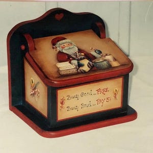 Puede incluir: Una caja decorativa de madera con una ilustración de Papá Noel en la tapa. La caja está pintada con colores rojo, negro y beige. La parte frontal de la caja tiene el texto "Beep Good...FREE, Beep Bad...PAY 5¢".