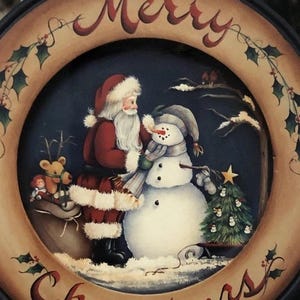 Puede incluir: Un plato decorativo redondo con temática navideña que presenta a Papá Noel y un muñeco de nieve. El plato tiene un borde marrón oscuro con las palabras "Merry Christmas" en escritura roja. La imagen central muestra a Papá Noel con un saco de juguetes y un muñeco de nieve.