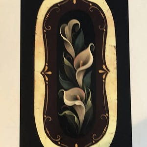 May include: A floral painting featuring three calla lilies in a dark brown oval frame with a cream-colored border. The lilies are white with a hint of pink, surrounded by green leaves, set against a black background. Decorative gold accents adorn the frame.