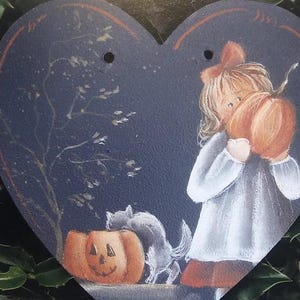 May include: Heart-shaped wooden decoration with a Halloween theme. It features a girl holding a pumpkin, a jack-o'-lantern, and a gray cat. The background is a dark blue with a tree silhouette. The decoration has two small holes at the top.