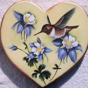 May include: A heart-shaped wooden plaque painted with a hummingbird in flight, surrounded by blue and white columbine flowers. The background is a pale yellow.