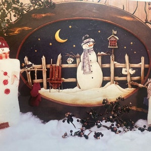 Puede incluir: Una escena invernal caprichosa con tres muñecos de nieve. El muñeco de nieve central lleva sombrero y bufanda, frente a una valla y un cielo nocturno con luna creciente y estrellas. Bayas decorativas y nieve completan la exhibición festiva.