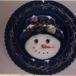 May include: A hand-painted ceramic plate with a snowman design. The snowman is wearing a black hat with a white trim and a red nose. The plate is decorated with a blue background and a vine border.