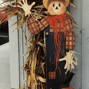 Puede incluir: Decoración de otoño de madera con un espantapájaros con sombrero, camisa a cuadros y peto. Un cuervo negro se posa sobre el fondo de tallos de maíz. Dos calabazas naranjas están en la base. El espantapájaros tiene una cara amigable y pelo de paja.