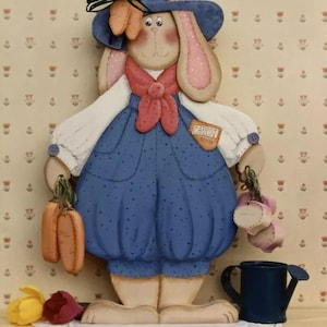 May include: A wooden bunny figurine wearing a blue hat, overalls, and a red scarf. The bunny is holding a bunch of carrots in one hand and a watering can in the other. The bunny is standing on a shelf with a white doily and a blue watering can.