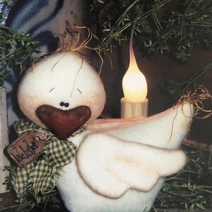 May include: A handcrafted, white fabric bird with a heart-shaped, burgundy beak and a green plaid scarf. The bird has a tag that reads "Webster". A battery-operated candle sits behind the bird, and the background includes greenery.