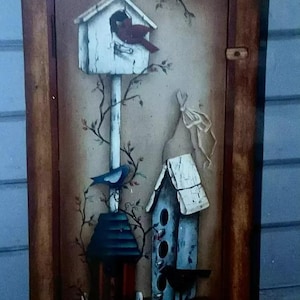 Puede incluir: Una puerta de armario de madera con una escena pintada de tres casas para pájaros. Las casas para pájaros están pintadas en tonos de azul, blanco y rojo. Un pájaro azul está posado en una de las casas para pájaros, y un cardenal está posado en otra. La puerta del armario está pintada en un patrón de veta de madera marrón.