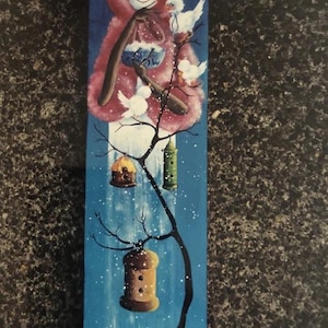 May include: Hand-painted wooden winter scene featuring a snowman in a red hooded coat, holding a blue bag, with white birds perched on a tree. The tree has birdhouses and a snowy mountain backdrop. The artwork is on a long, narrow wooden board.