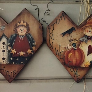 Puede incluir: Dos adornos de pared de madera en forma de corazón. Uno presenta una figura con corona, estrellas y una bandera estadounidense. El otro representa un espantapájaros con una calabaza y un cuervo. Ambos tienen la palabra "WELCOME".