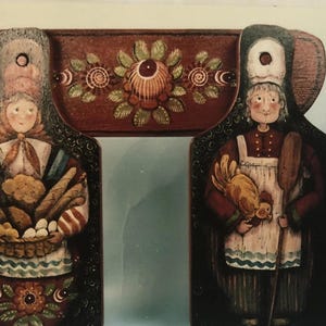 May include: Wooden wall decor featuring two figures in traditional attire. One figure holds a basket of bread and eggs, the other holds a chicken and a wooden spoon. The figures are connected by a decorative wooden arch.