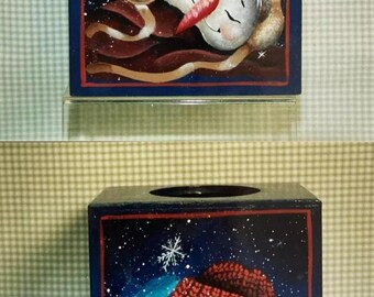 Puede incluir: Dos cubiertas de caja de pañuelos de madera con un tema invernal. La cubierta superior presenta un muñeco de nieve con una nariz roja y un sombrero marrón durmiendo sobre un fondo azul con copos de nieve blancos. La cubierta inferior presenta un muñeco de nieve con un sombrero rojo y una bufanda verde durmiendo sobre un fondo azul con copos de nieve blancos.