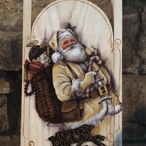 May include: Wooden Christmas sled with a painted image of Santa Claus holding a candy cane and a sack of toys. The sled has the words "MERRY CHRISTMAS" in a decorative font.
