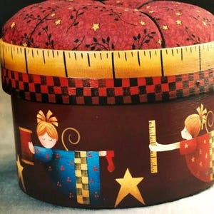 May include: A round, red and white fabric-covered box with a yellow measuring tape around the top. The box is decorated with painted figures of two children, one holding a red ribbon and the other holding a ruler. The box is decorated with a black and white checkered pattern and gold stars.