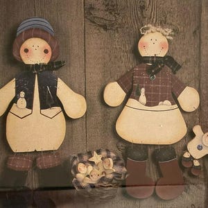 May include: Two wooden snowman figures with painted details. The male snowman is wearing a blue hat, a black vest, and brown boots. The female snowman is wearing a plaid dress and brown boots. Both snowmen are holding small white snowballs. A basket of smaller snowmen is in the foreground.