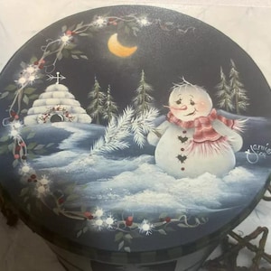 Könnte beinhalten: Runde, handbemalte dekorative Box mit einer Winterszene. Ein lächelnder Schneemann mit einem rot-weiß gestreiften Schal steht im Schnee. Der Hintergrund zeigt ein Iglu, Kiefern und einen Halbmond. Die Box ist mit einem dekorativen Rand versehen.