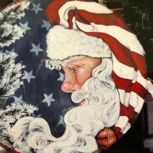May include: A painted wooden round ornament featuring a Santa Claus with a white beard and red, white, and blue striped hat. The background is a blue and white American flag.