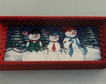 Puede incluir: Una bandeja rectangular de madera roja con asas recortadas. El interior de la bandeja presenta una escena invernal con tres muñecos de nieve con sombreros, bufandas y corbatas. El fondo muestra árboles cubiertos de nieve y una valla.