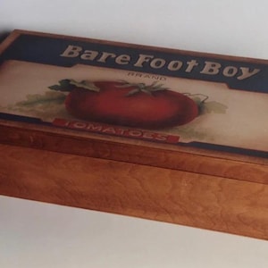 Può includere: Una scatola rettangolare in legno con coperchio incernierato. Il coperchio presenta un disegno dipinto con le parole "Bare Foot Boy BRAND TOMATOES" e una grande illustrazione di pomodoro rosso. La scatola ha una finitura naturale.