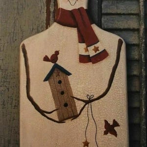 May include: A snowman-shaped decorative wooden plaque with a red scarf and hat, and a carrot nose. The plaque features a birdhouse, birds, and star accents, with a distressed, crackled finish. The overall theme is winter and holiday decor.