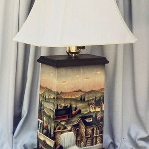 Puede incluir: Una lámpara de mesa con una pantalla blanca y una base cuadrada. La base está pintada con una escena de un pueblo pequeño con una cascada y una rueda de agua.