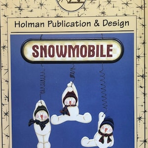 May include: A blue and cream-colored print featuring the text "SNOWMOBILE" above three hanging snowman figures. Each snowman wears a hat and scarf. The print is from Holman Publication & Design.
