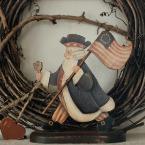 Puede incluir: Figura de madera del Tío Sam, pintada en rojo, blanco y azul, sosteniendo una bandera y un pequeño objeto. La figura está colocada frente a una corona de mimbre. Adornos en forma de corazón en rojo y azul en primer plano.