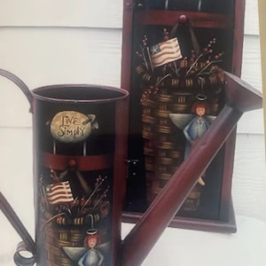 Puede incluir: Una regadera roja pintada con un asa y un pico. La regadera está decorada con una escena pintada de una cesta de flores, una bandera estadounidense y un ángel. El texto "Live Simply" está pintado en la regadera.