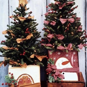 Könnte beinhalten: Zwei kleine Weihnachtsbäume in dekorativen Kisten. Eine Kiste zeigt einen Lebkuchenmann, die andere einen Schneemann. Beide Bäume sind mit Schleifen, Beeren und Stern- oder Herzaufsätzen geschmückt. Die Kisten sind in festlichen Farben bemalt.